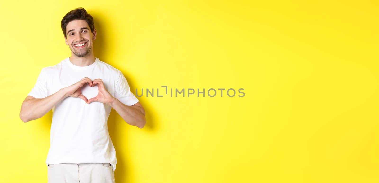 Happy romantic man showing heart sign, smiling and express love, standing over yellow background. Copy space