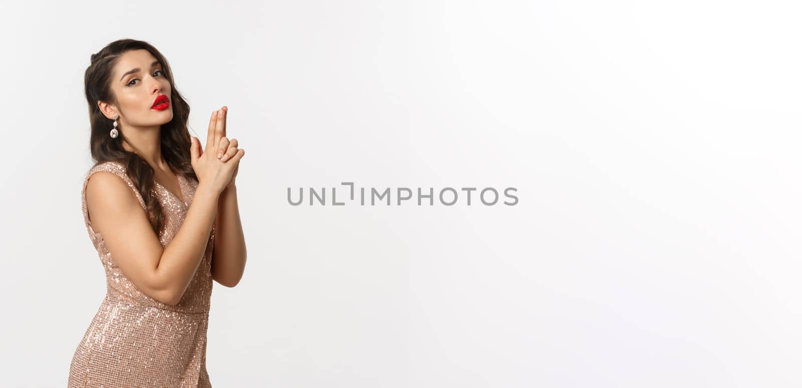 Concept of casino, celebration and party. Sexy woman in glamour dress, red lips, making finger gun pistol gesture and looking at camera, standing over white background.