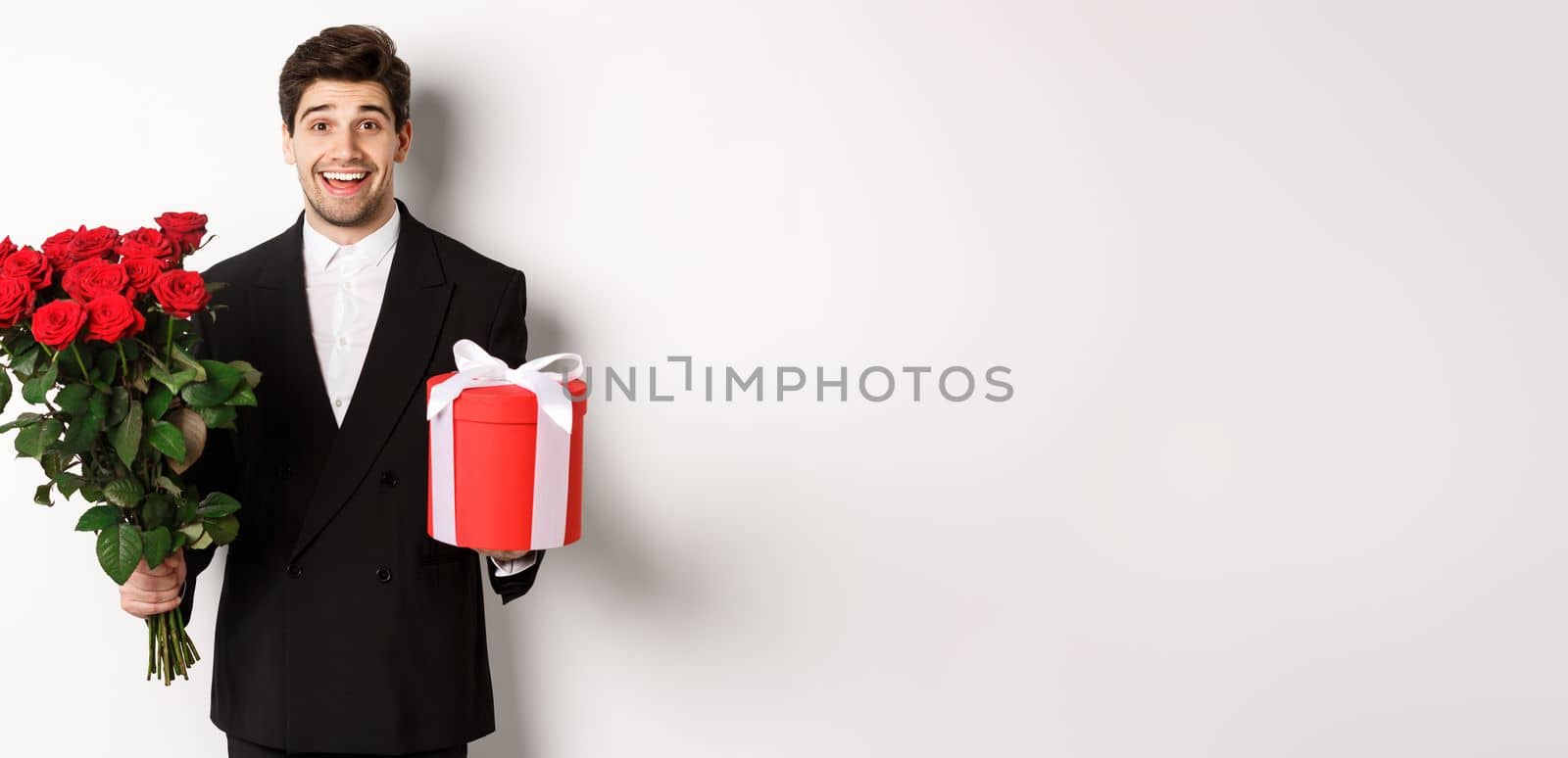 Concept of holidays, relationship and celebration. Image of handsome smiling guy in black suit, holding bouquet of red roses and giving present on new year, white background by Benzoix