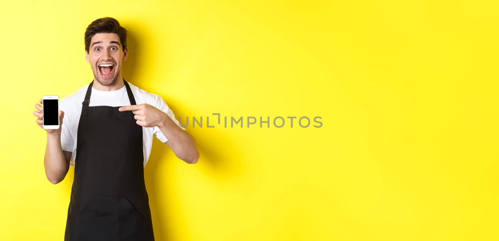 Handsome barista in black apron pointing finger at mobile screen, showing app and smiling, standing over yellow background by Benzoix