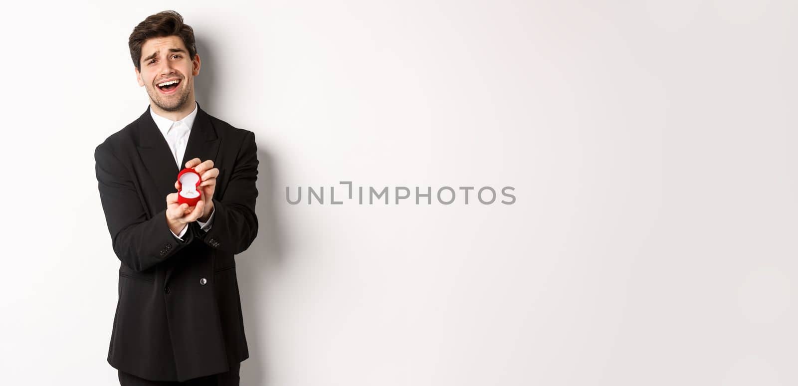 Portrait of handsome man in black suit, open box with wedding ring, making a proposal, asking to marry him, standing against white background.