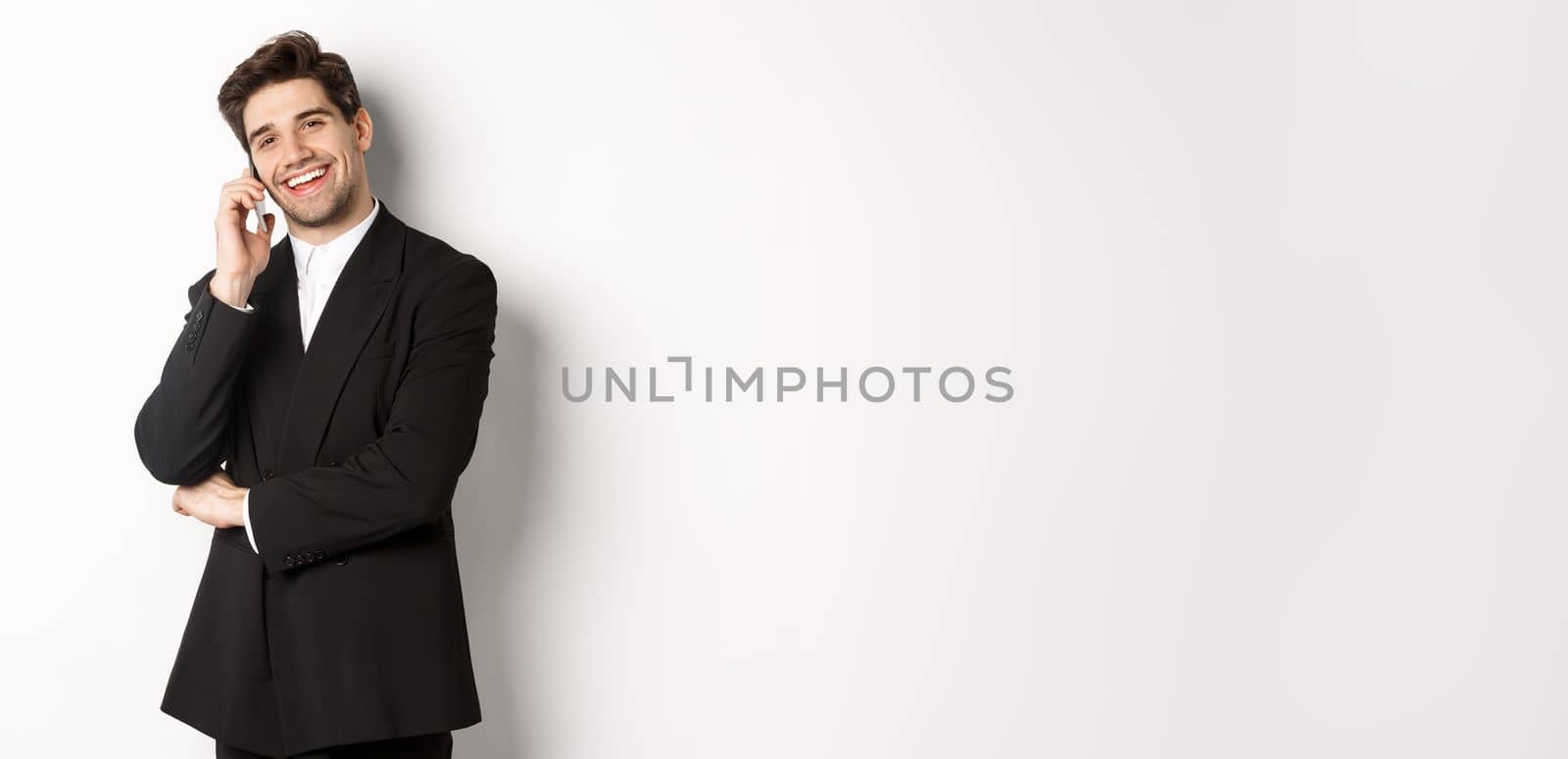 Image of handsome, successful businessman talking on phone, smiling pleased, standing in suit against white background.