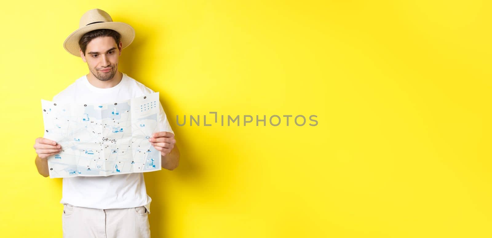 Travelling, vacation and tourism concept. Male tourist looking confused at map, looking for right direction, standing over yellow background.