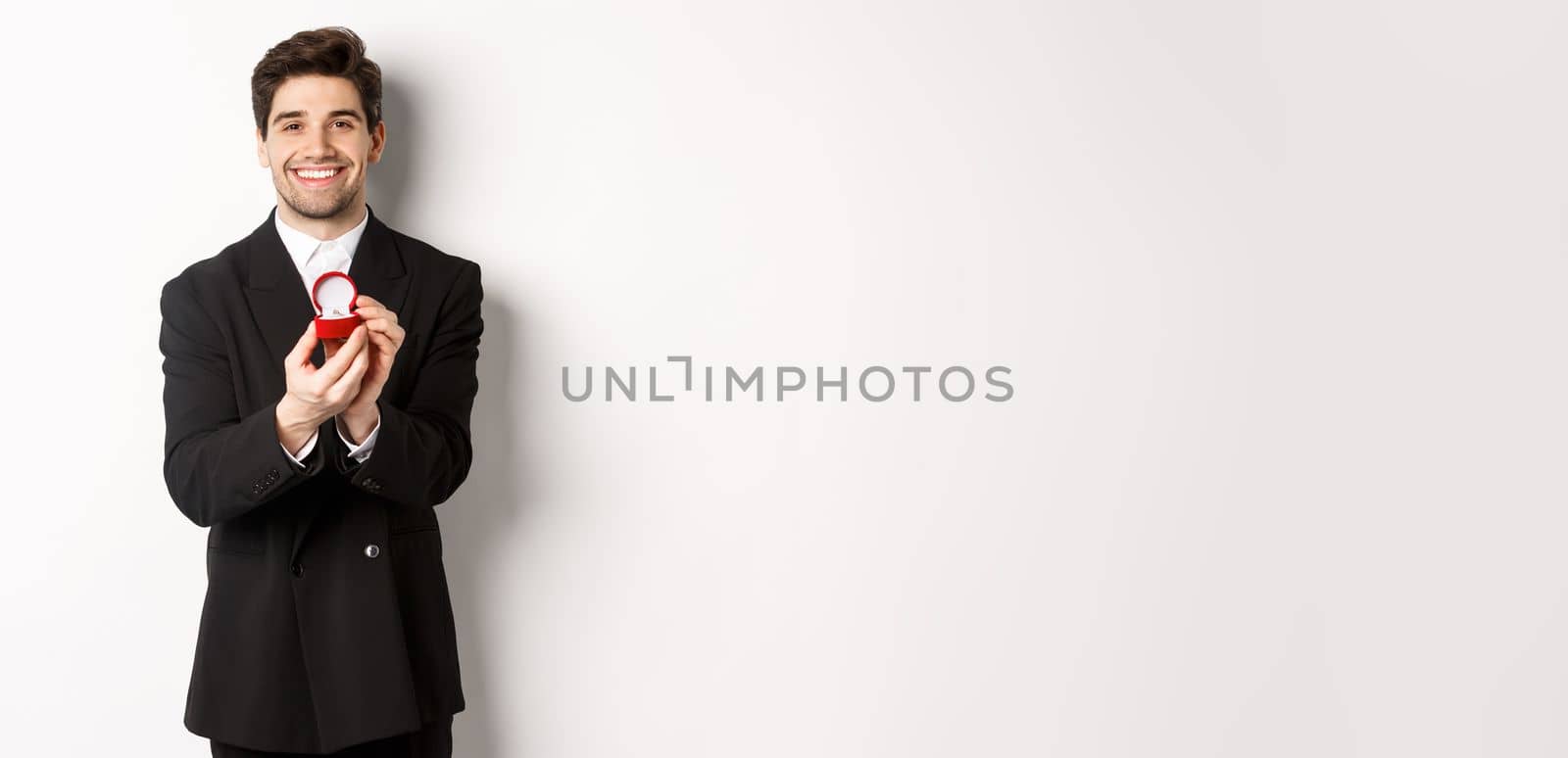 Image of handsome boyfriend in black suit making a proposal, asking to marry him and showing wedding ring, standing over white background.