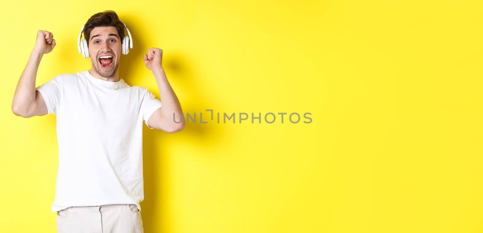 Excited handsome man dancing and singing along, listening music in headphones, standing over yellow background.