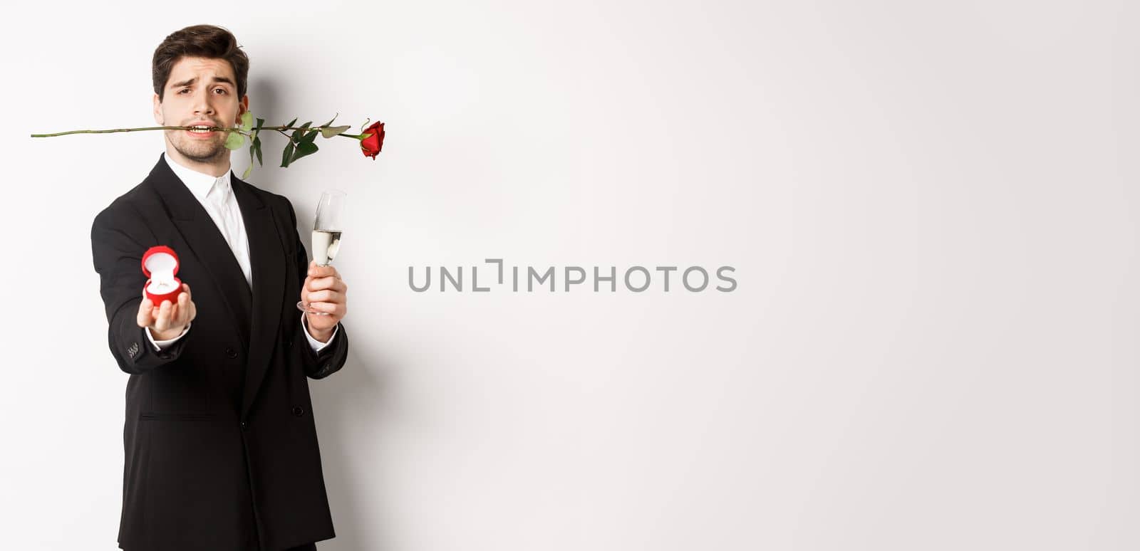 Romantic young man in suit making a proposal, holding rose in teeth and glass of champagne, showing engagement ring, asking to marry him, standing against white background.