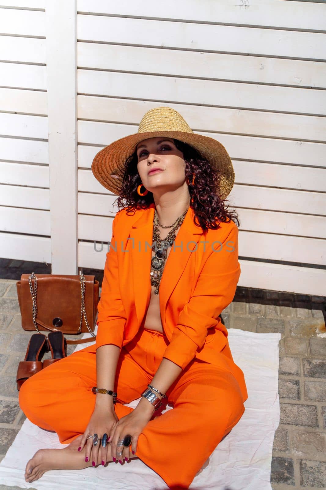 Stylish woman in an orange suit with a hat sits on a rug on a white striped background. On the hands are jewelry rings and bracelets, sandals and a bag stand side by side