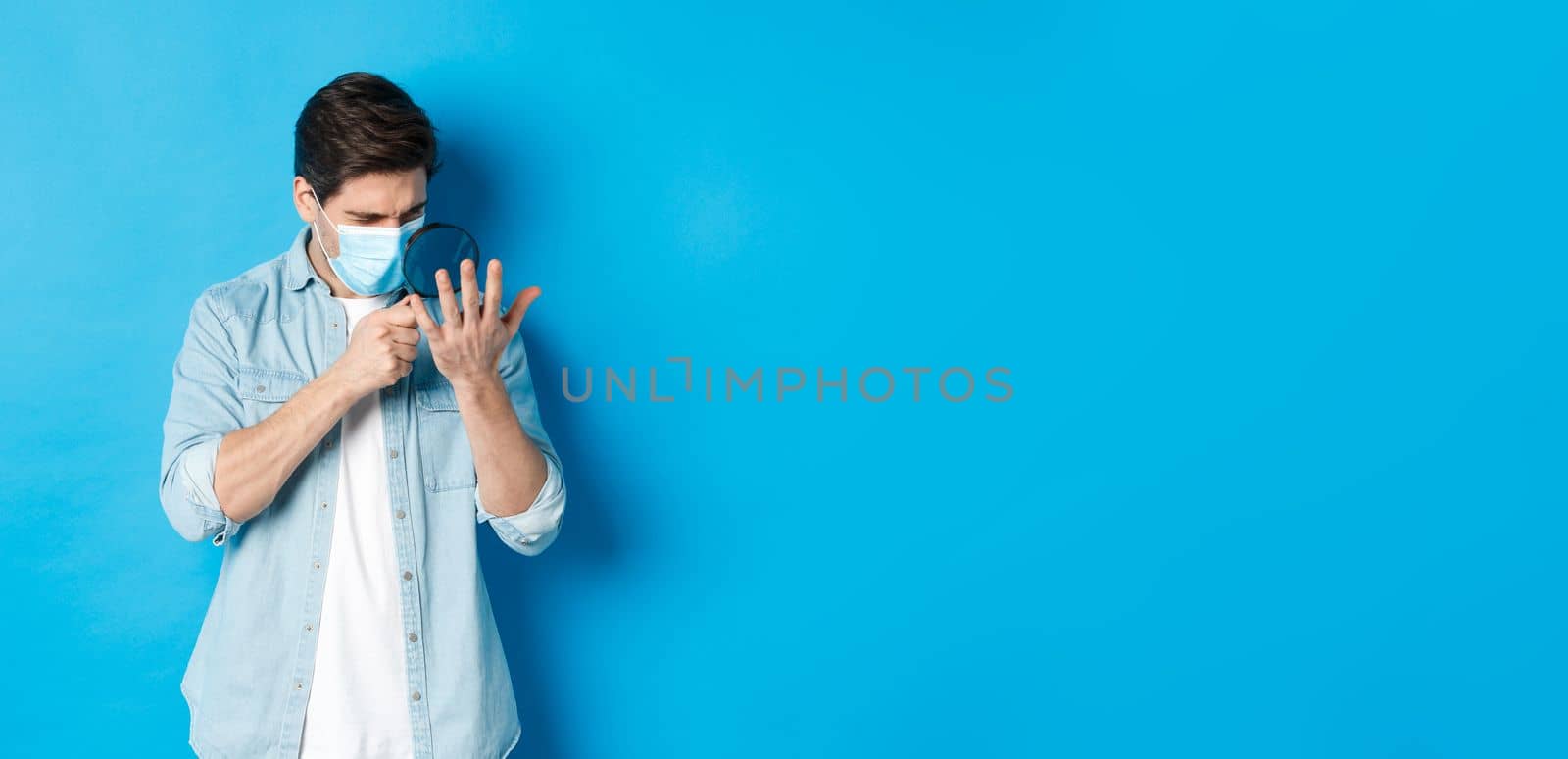 Concept of coronavirus, social distancing and pandemic. Man in medical mask looking at his palm through magnifying glass, standing over blue background by Benzoix