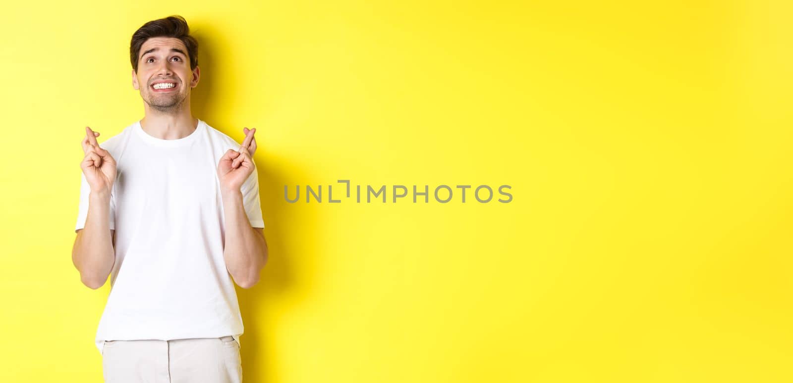 Nervous and hopeful man praying to god, making wish with fingers crossed, panicking and standing over yellow background.