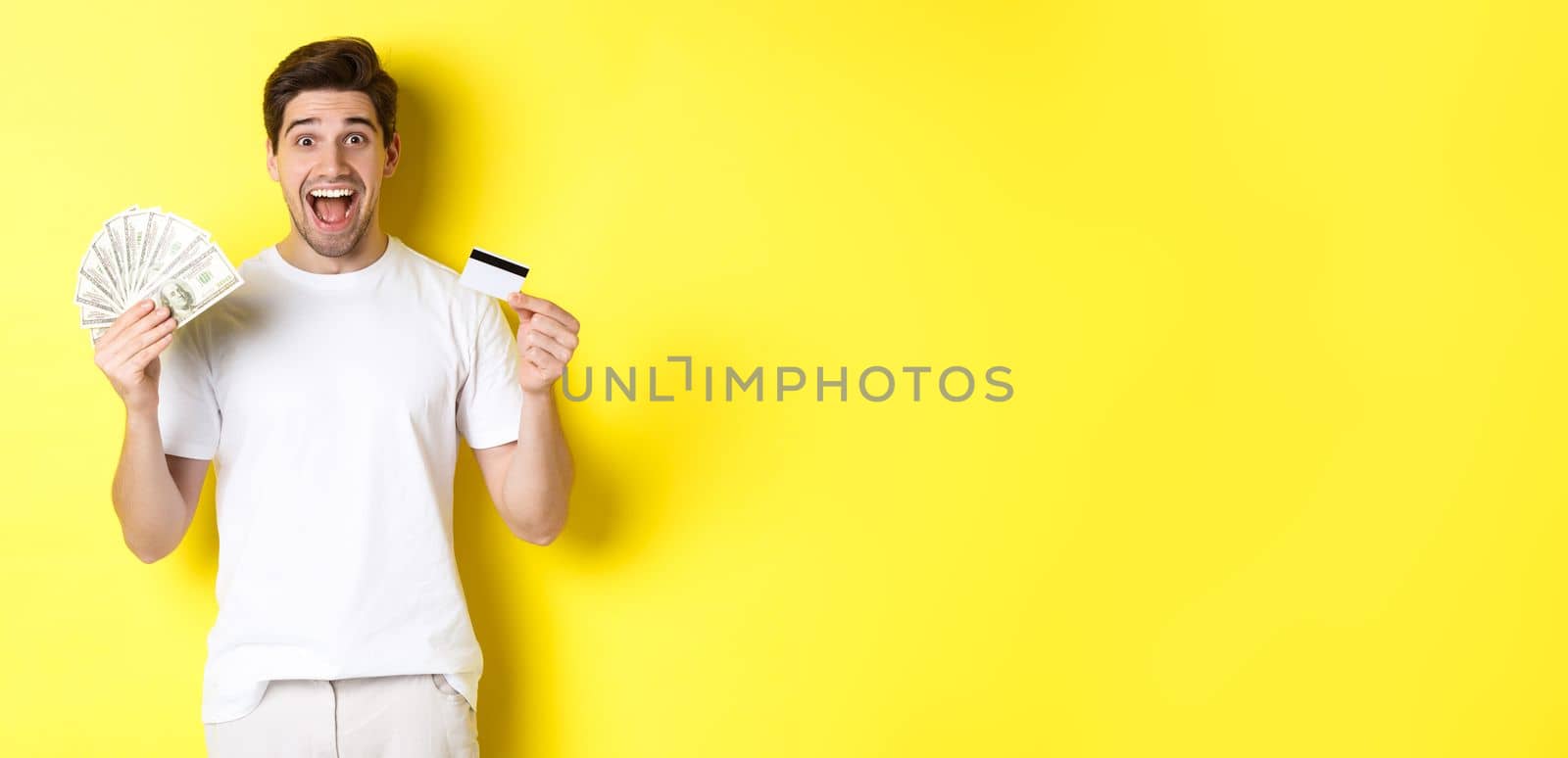 Excited man ready for black friday shopping, holding money and credit card, standing over yellow background.