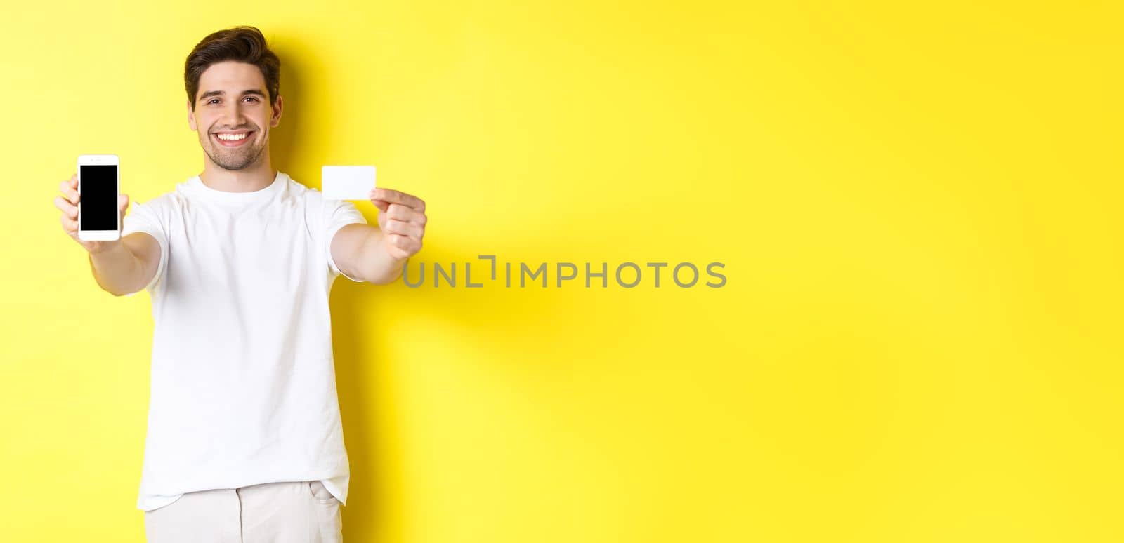 Handsome caucasian male model showing smartphone screen and credit card, concept of mobile banking and online shopping, yellow background by Benzoix