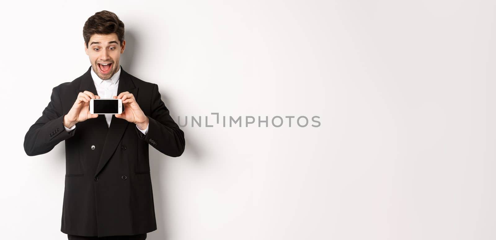 Portrait of excited handsome man showing smartphone screen, looking amazed at display with advertisement, standing over white background in black suit.