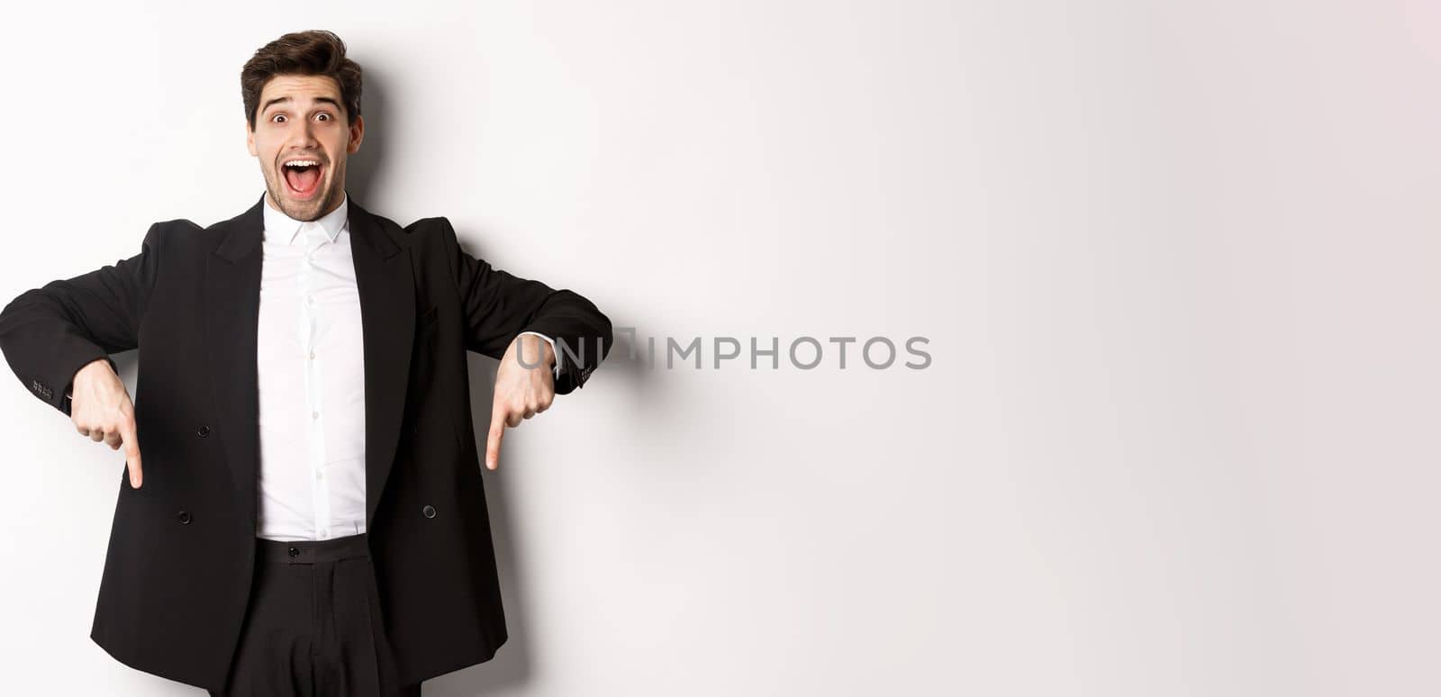 Portrait of super happy man showing christmas promo offer, pointing fingers down and smiling amazed, wearing formal party suit, standing over white background.