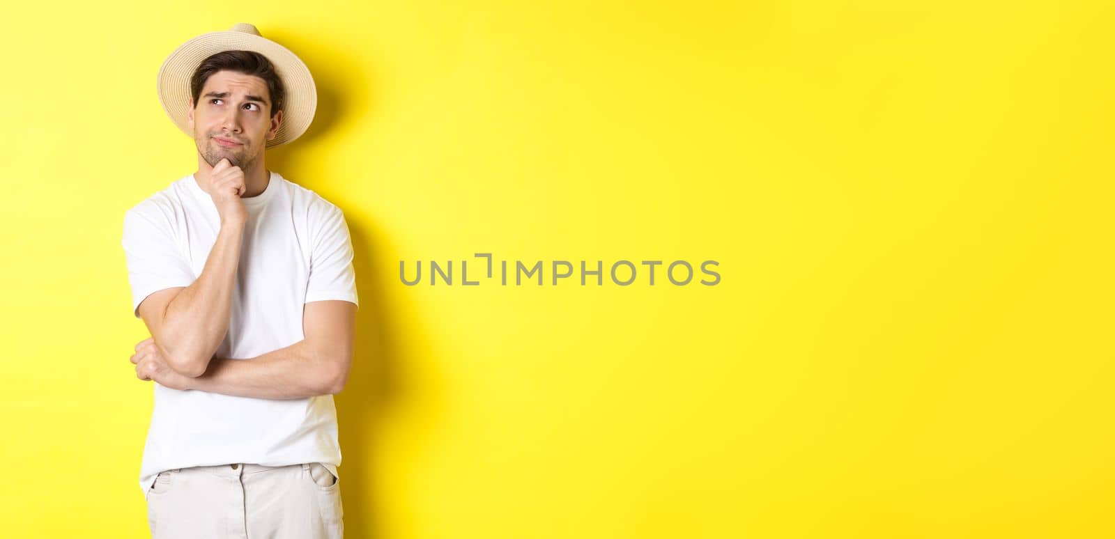 Concept of tourism and summer. Thoughtful man tourist pondering, looking at upper left corner and thinking, standing in straw hat and white t-shirt against yellow background.