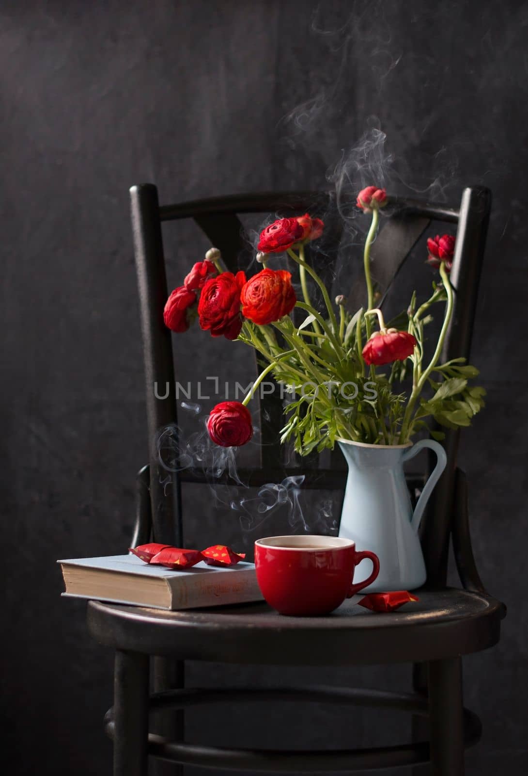 vintage still life on a black chair against a black background, red ranunculi in a blue jug, a red cup of tea with smoke, a blue book and candies in a red wrapper, dark photo, High quality photo