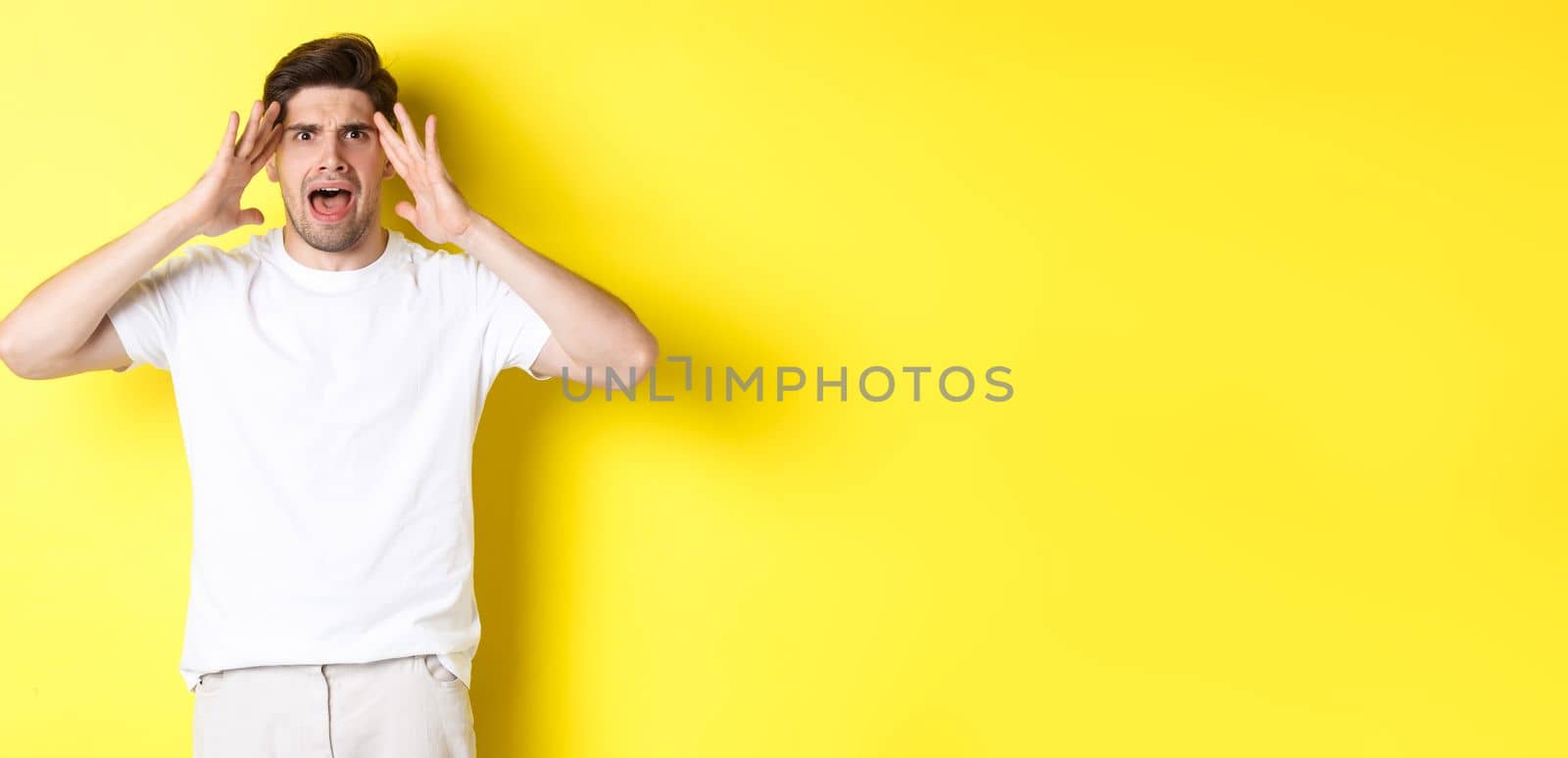 Frustrated guy looking alarmed, holding hands on head and feeling confused, panicking, standing over yellow background.