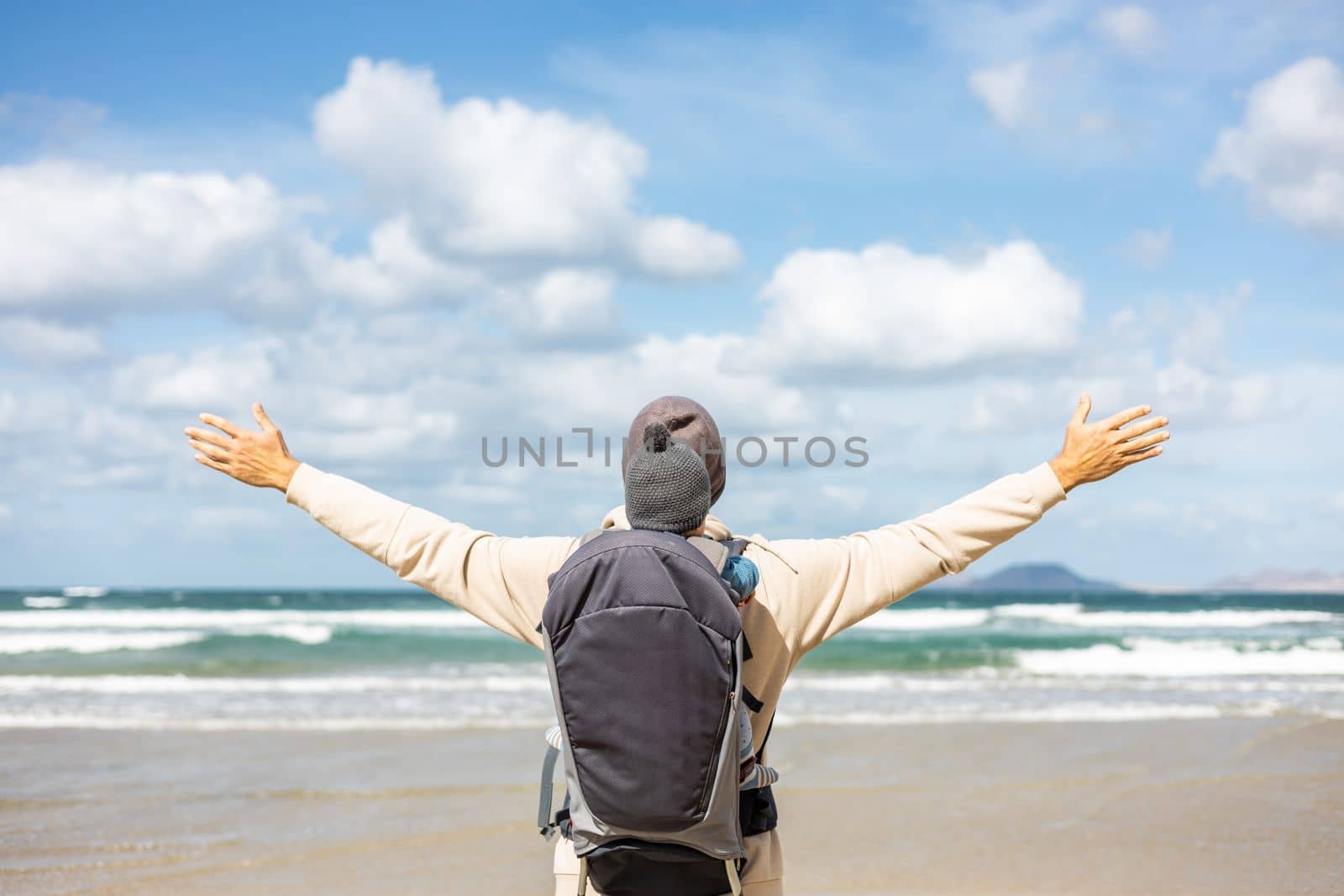 Young father rising hands to the sky while enjoying pure nature carrying his infant baby boy son in backpack on windy sandy beach of Famara, Lanzarote island, Spain. Family travel concept. by kasto
