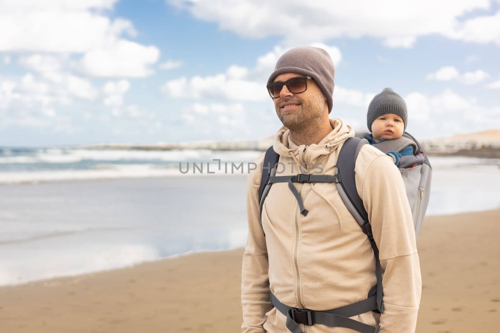 Young father carrying hisinf ant baby boy son in backpack on windy sandy Famara beach, Lanzarote island, Spain. Family travel and winter vacation concept