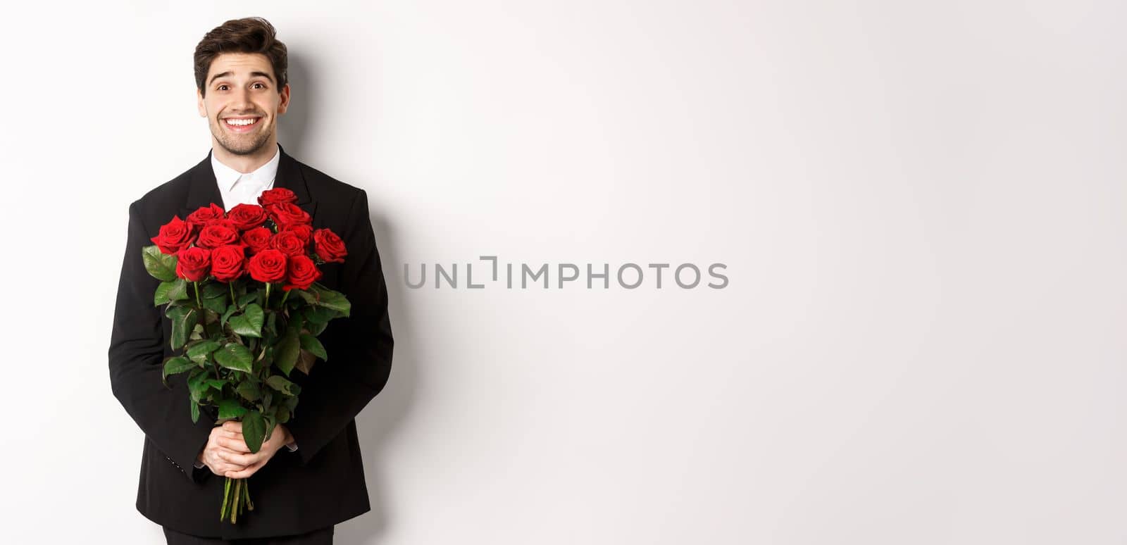 Image of handsome man in black suit, holding bouquet of roses and smiling, standing against white background.