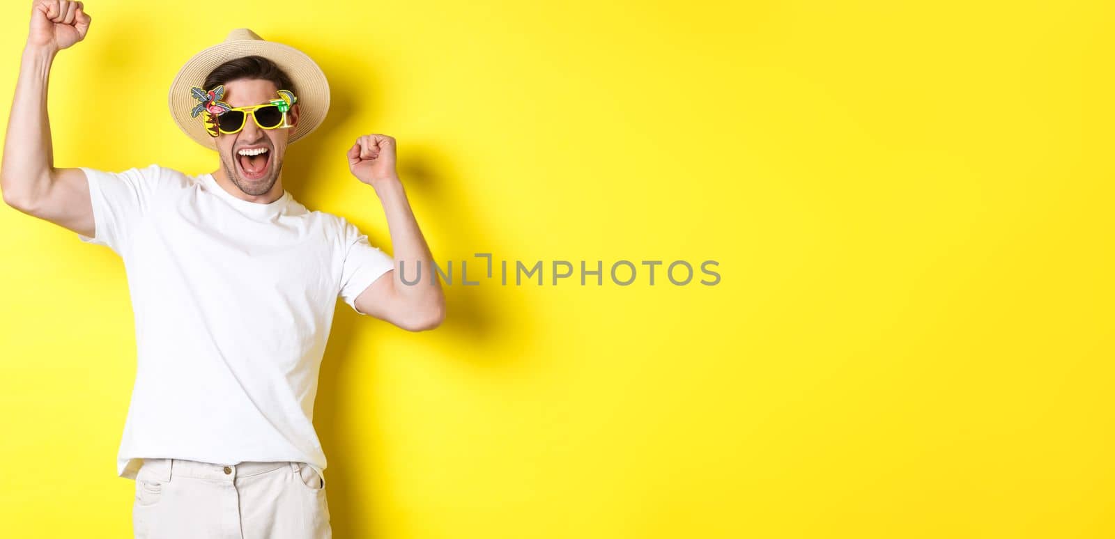 Concept of tourism and lifestyle. Happy lucky guy winning trip, rejoicing and wearing holiday outfit, summer hat and sunglasses, yellow background by Benzoix