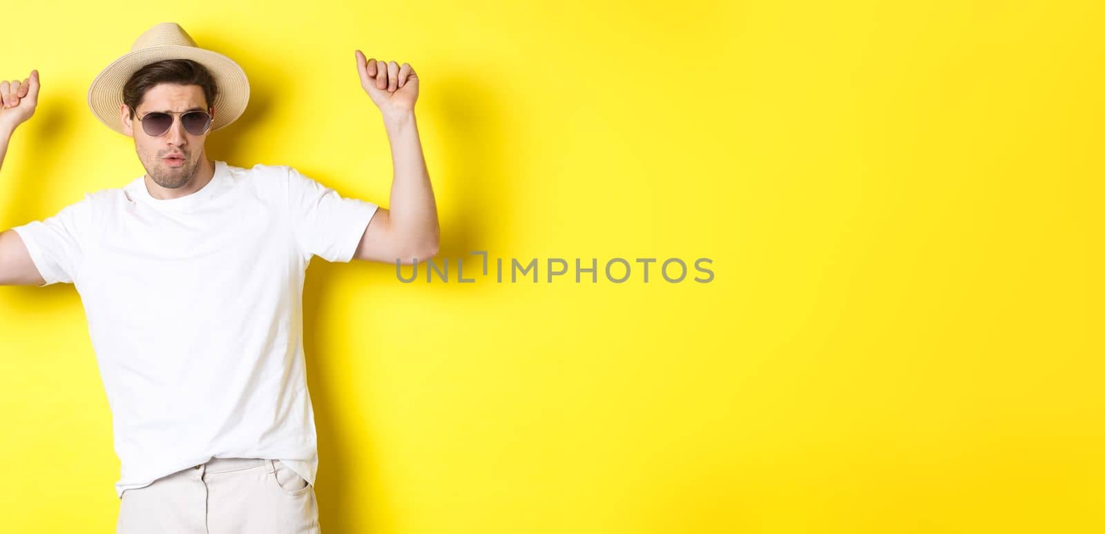 Tourism, travelling and holidays concept. Man tourist enjoying vacation, dancing in straw hat and sunglasses, posing against yellow background.