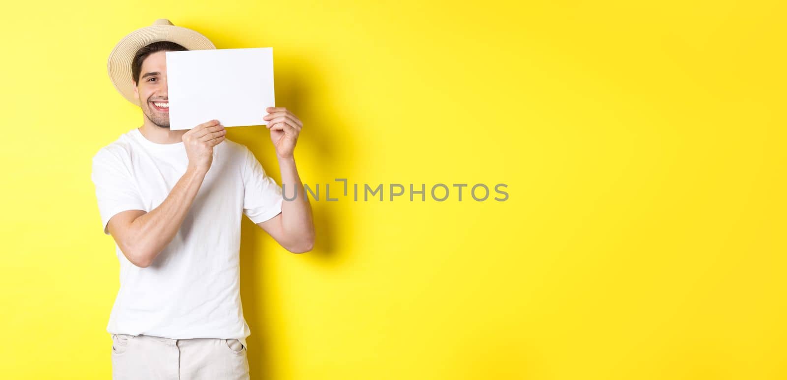 Happy guy on vacation showing blank piece of paper for your logo, holding sign near face and smiling, standing against yellow background.