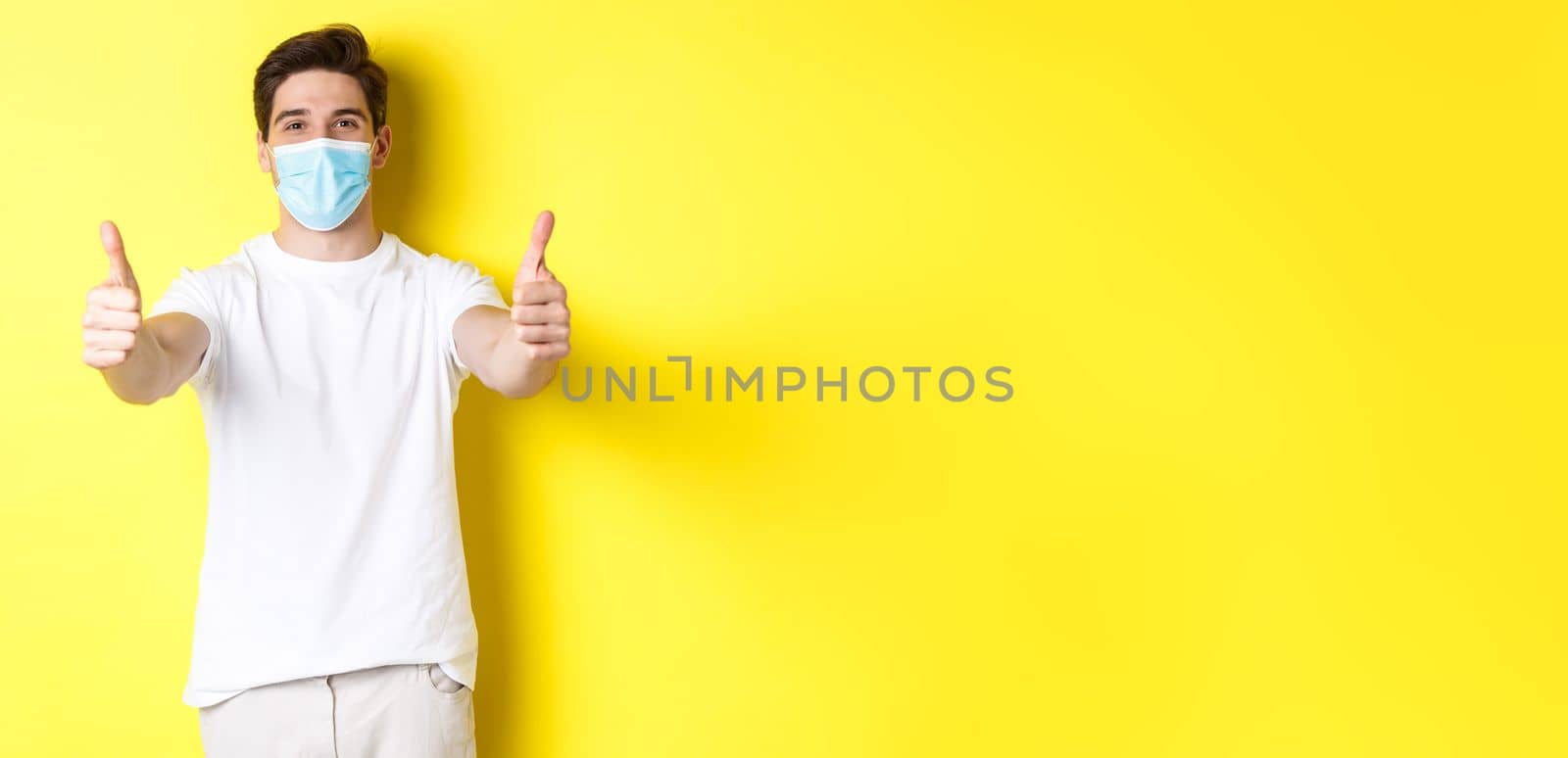 Concept of coronavirus, pandemic and social distancing. Confident man protecting himself from covid-19 with medical mask, showing thumbs up in approval, yellow background by Benzoix