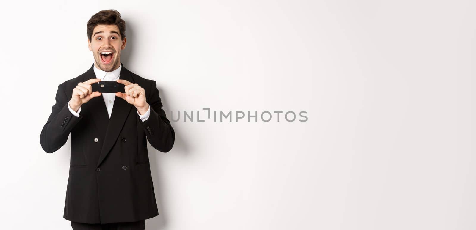 Portrait of amazed handsome man in black suit, showing credit card and recommending bank, standing over white background.