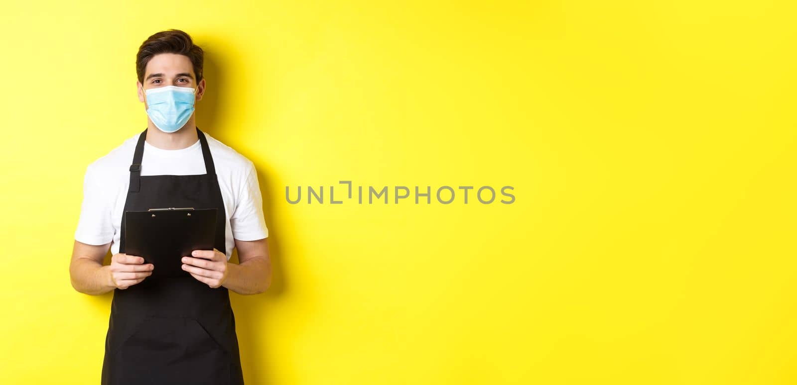 Concept of covid-19, small business and quarantine. Salesman in black apron and medical mask holding clipboard, working in shop, standing over yellow background by Benzoix