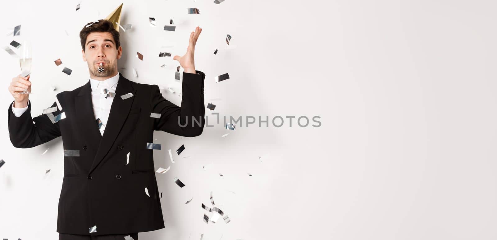 Handsome party guy in black suit having fun, celebrating new year, blowing whistle and drinking champagne while confetti falling, wishing happy holidays, standing against white background.