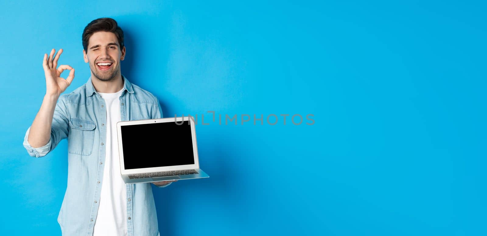 Young man showing laptop screen and okay sign, approve or like promo in internet, smiling satisfied, standing over blue background by Benzoix