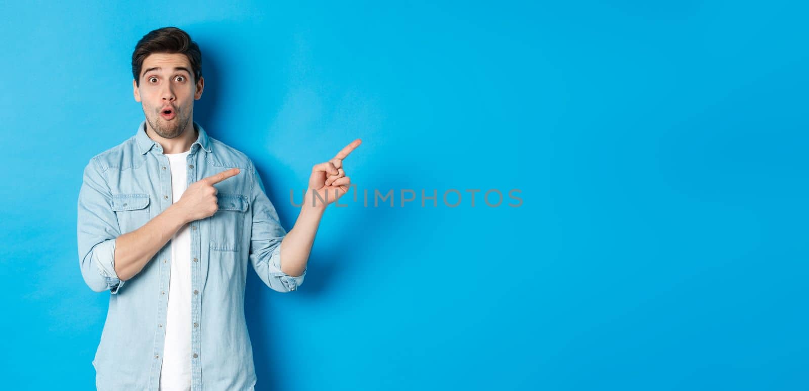 Portrait of surprised bearded guy in casual outfit pointing fingers right, showing amazing promo offer, standing over blue background.