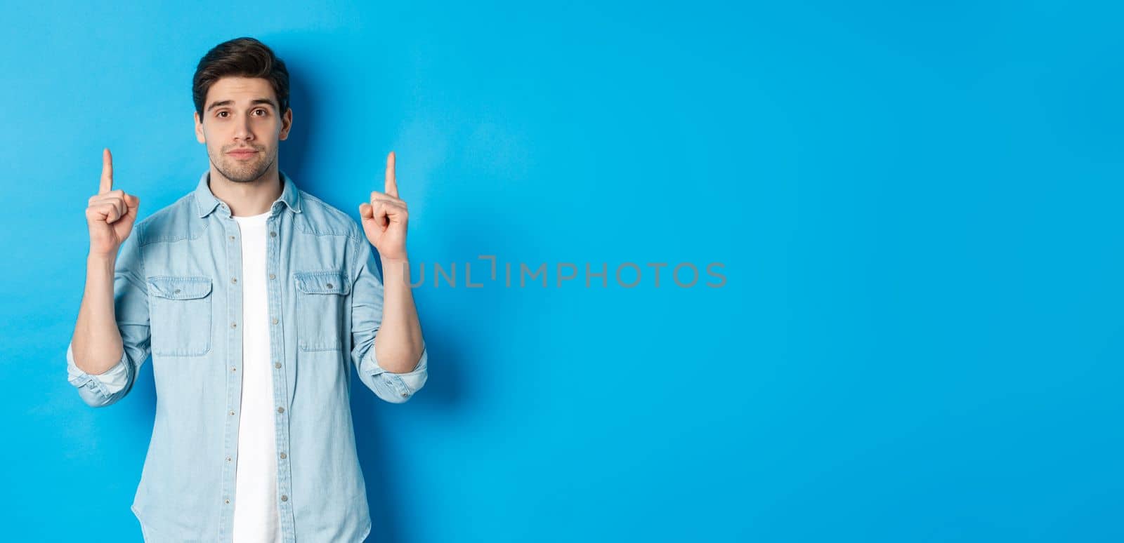 Image of calm handsome man showing you promo offer, pointing fingers up at copy space, standing against blue background.