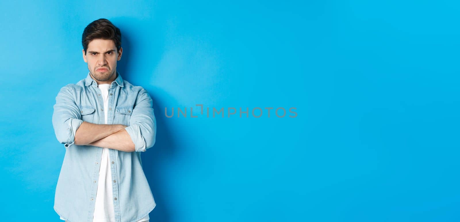 Angry bearded guy crossing arms on chest, frowning, looking mad and offended, standing against blue background.