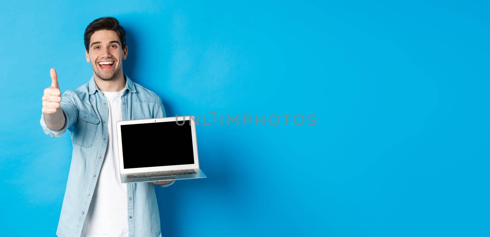 Image of satisfied handsome man showing laptop screen, thumbs-up in approval, like website or internet, standing over blue background.