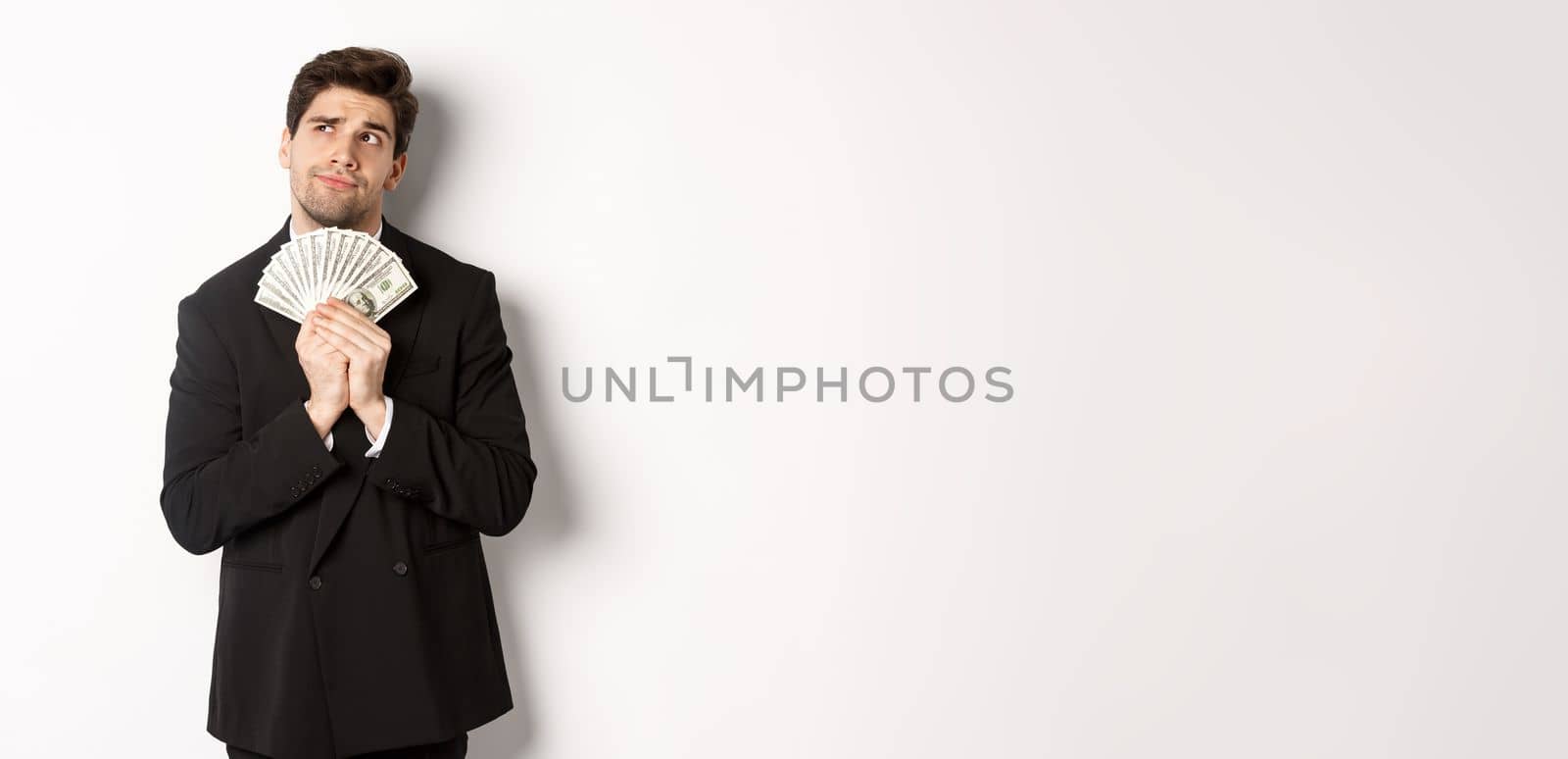 Image of handsome businessman in black suit, holding money and thinking about investment, making shopping plans, standing over white background.