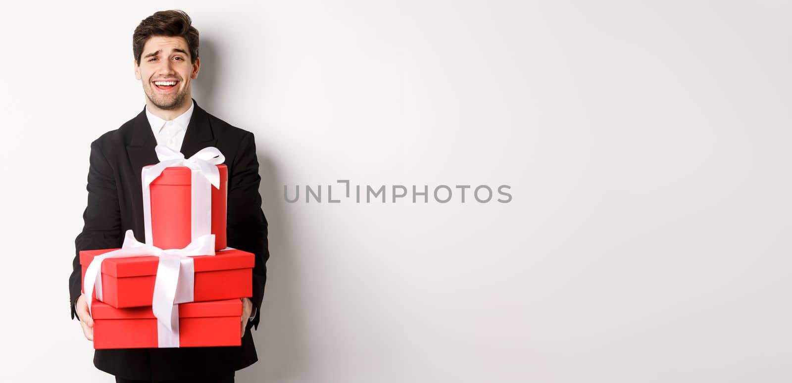 Image of handsome guy in black suit, holding gifts for christmas holidays, standing against white background.