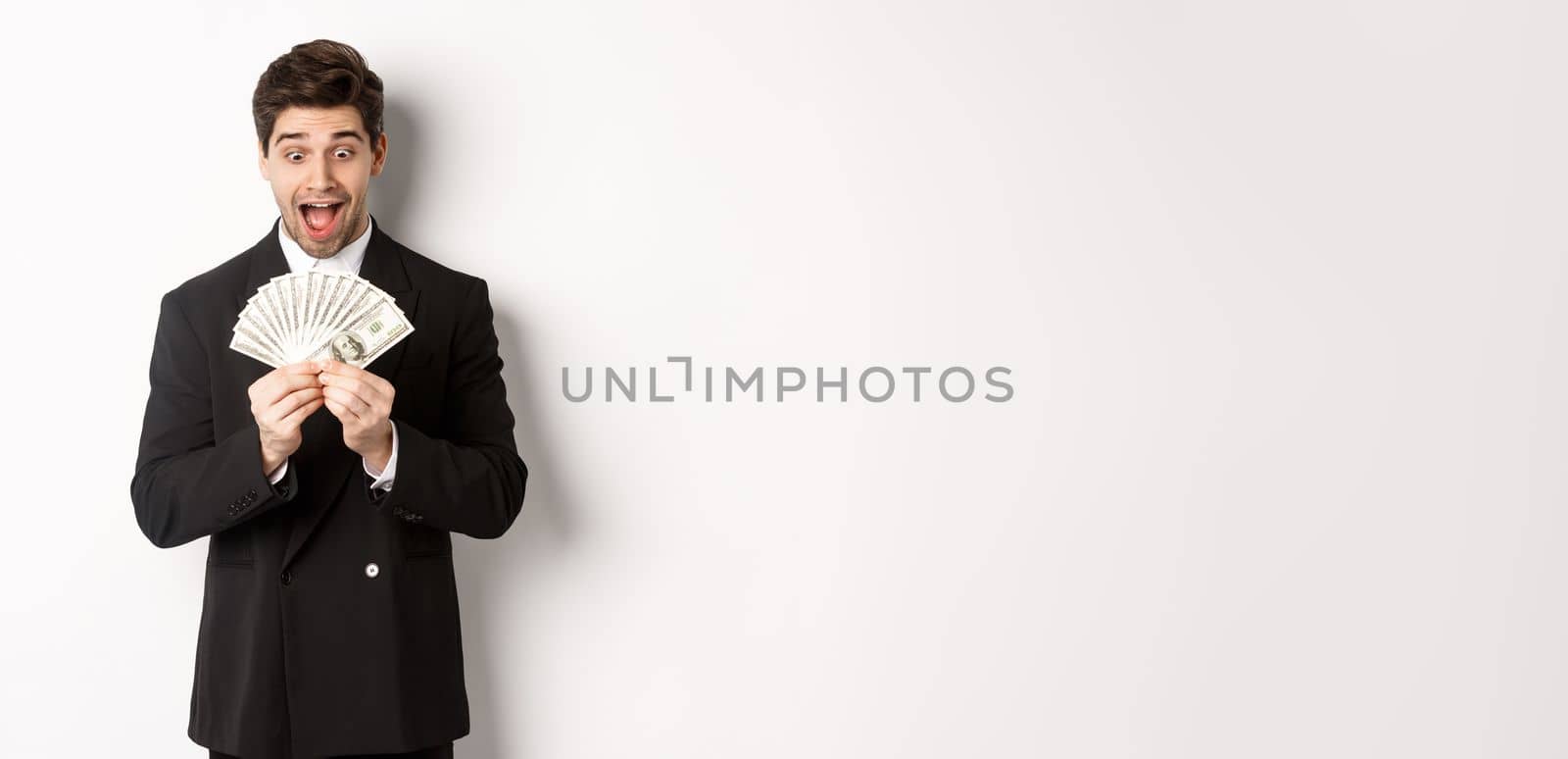 Image of handsome bearded guy in black suit, looking at money with excitement, standing over white background.