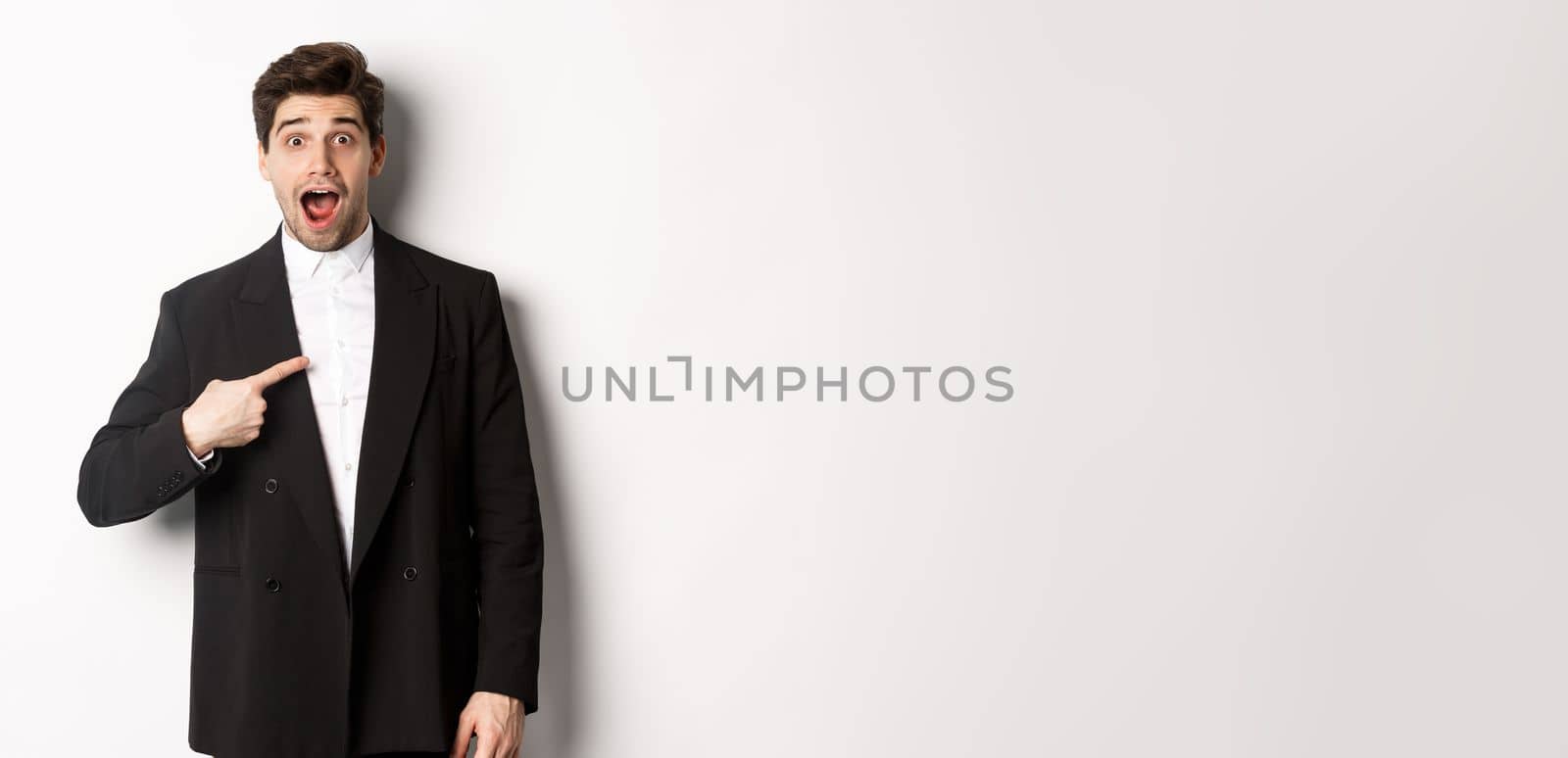 Concept of new year party, celebration and lifestyle. Portrait of surprised handsome guy in black suit, pointing at himself and looking amazed, being chosen, standing over white background.