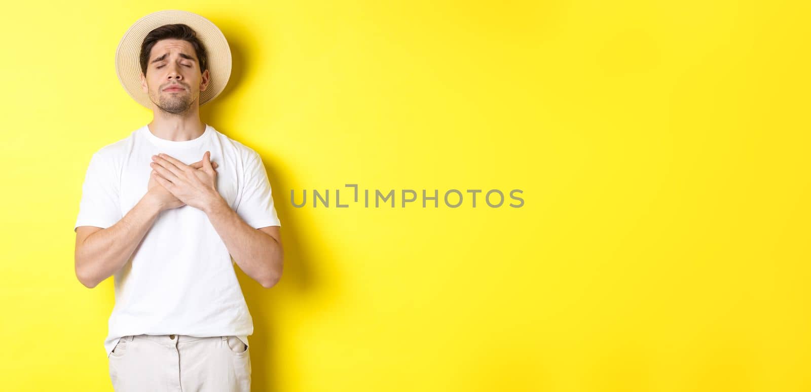 Concept of tourism and summer. Romantic man in straw hat looking nostalgic, close eyes and holding hands on heart, standing against yellow background.