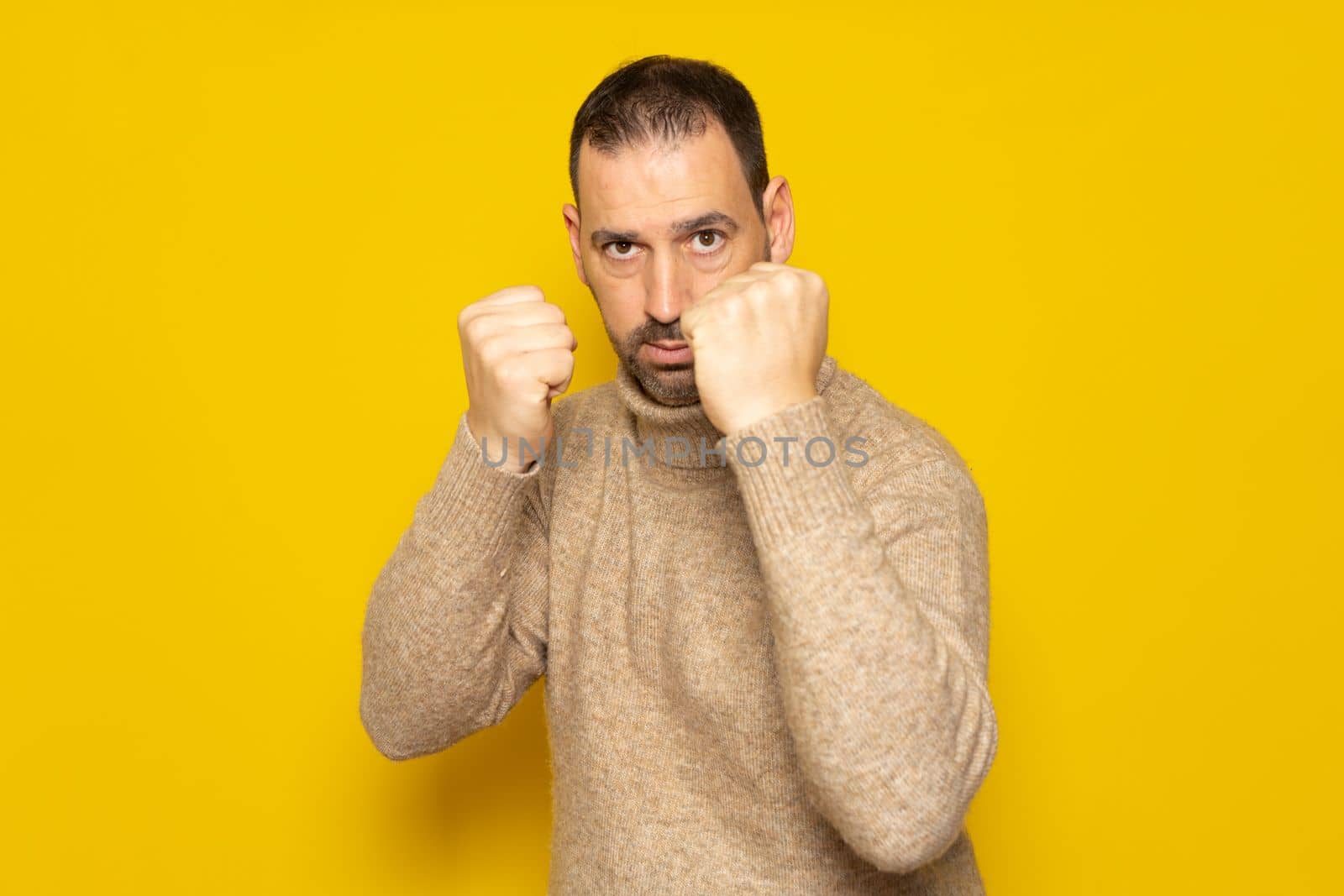 Handsome bearded man wearing turtleneck sweater over yellow background. Banging fist to fight, aggressive and angry attack, threat and violence