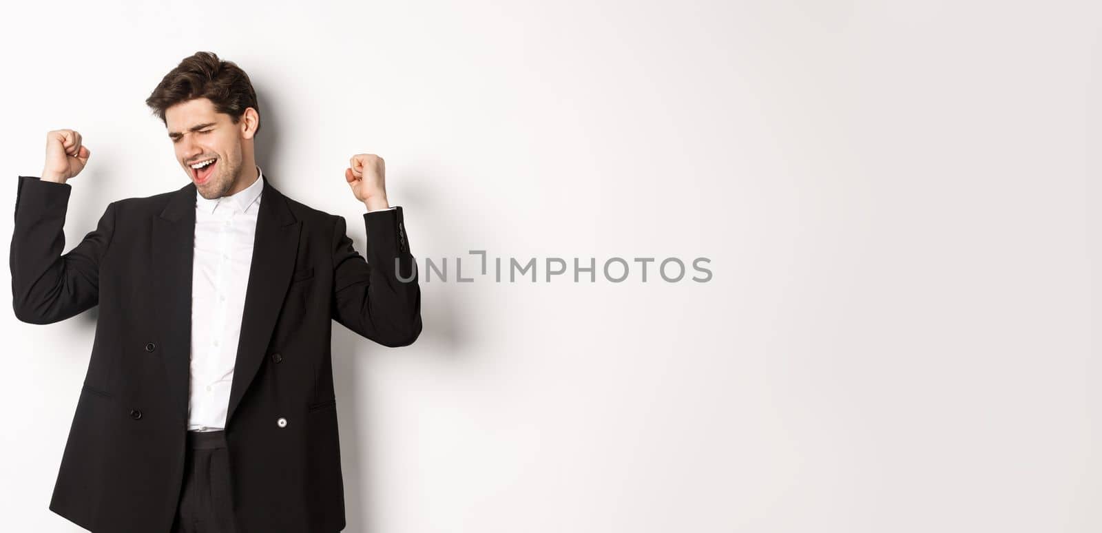 Image of successful and pleased handsome man in suit, rejoicing and making fist pump signs, dancing from happiness, standing over white background by Benzoix