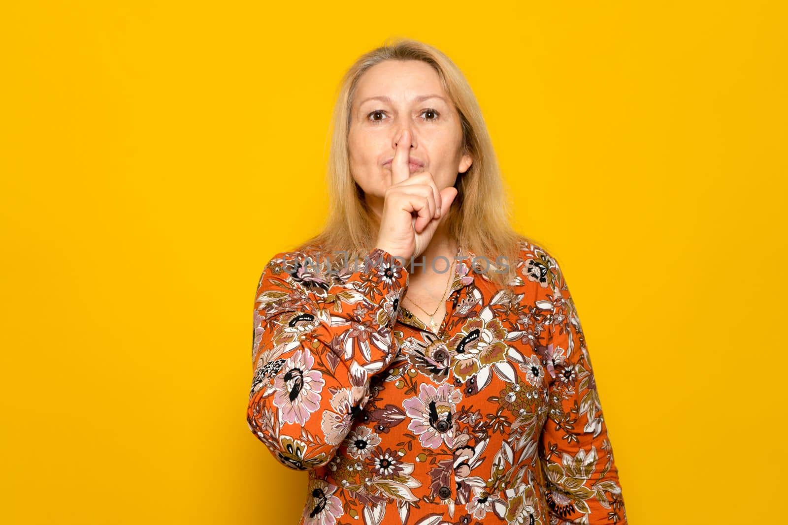 Pretty caucasian blonde woman in a patterned dress making the silence gesture with her index finger. She wants to create an aura of mystery, she hides a secret