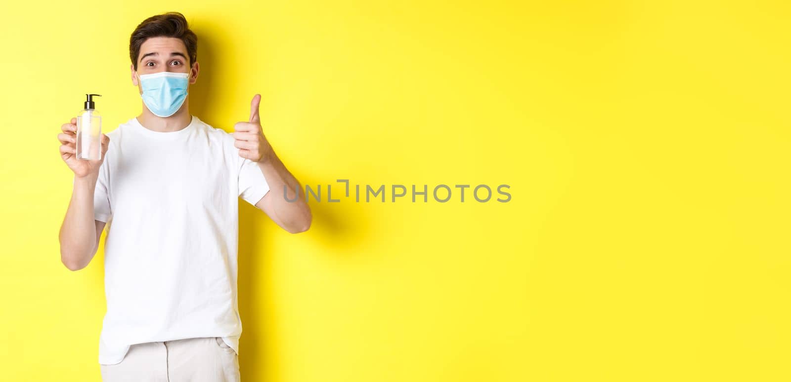 Concept of covid-19, quarantine and lifestyle. Satisfied young man in medical mask showing good hand sanitizer, thumbs up and recommending antiseptic, yellow background by Benzoix