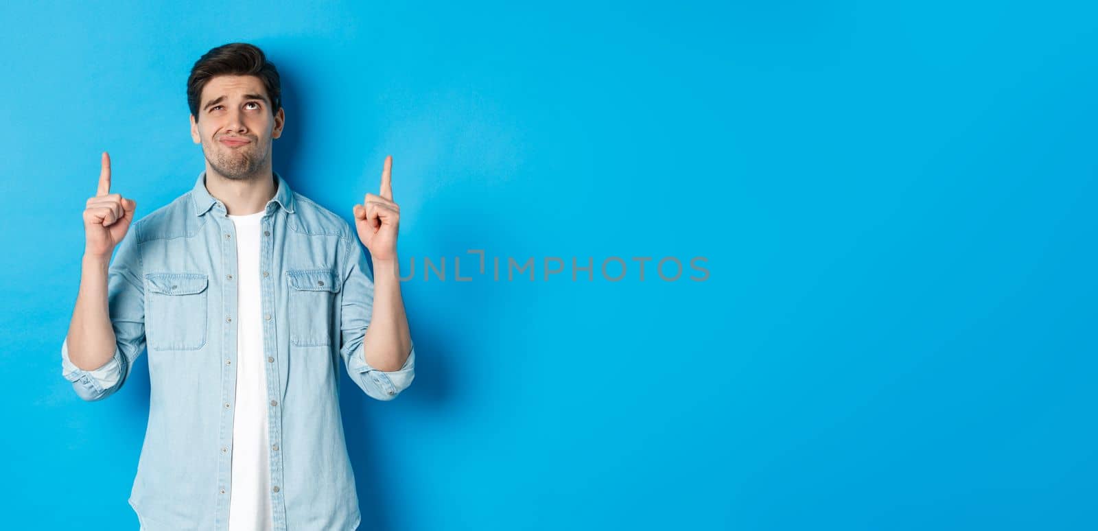Portrait of doubtful adult man looking disappointed, pointing fingers up at something with uncertain face, standing over blue background.