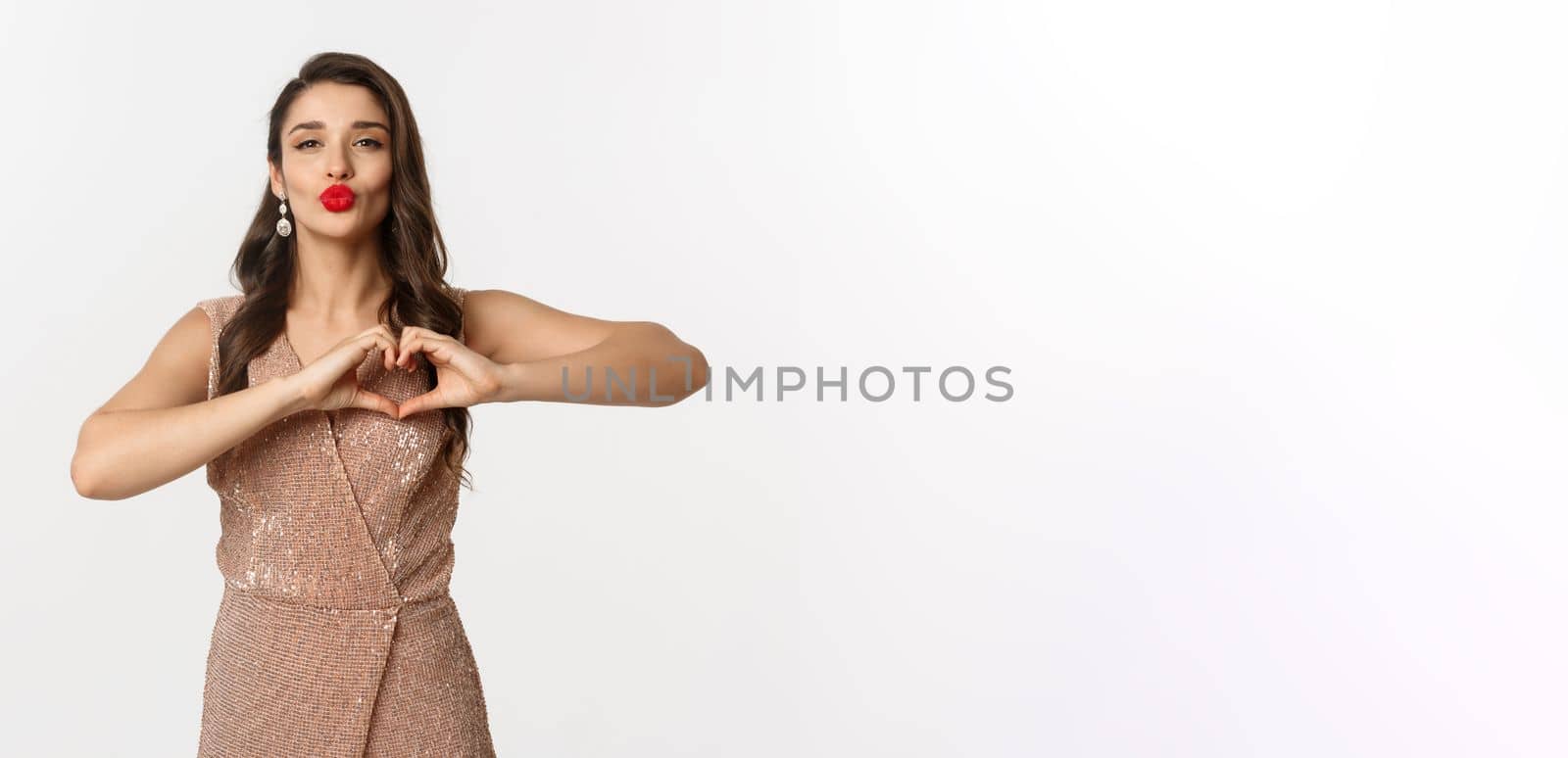 Celebration and party concept. Beautiful young woman in stylish dress, red lips, showing heart sign and waiting for kiss, standing over white background by Benzoix