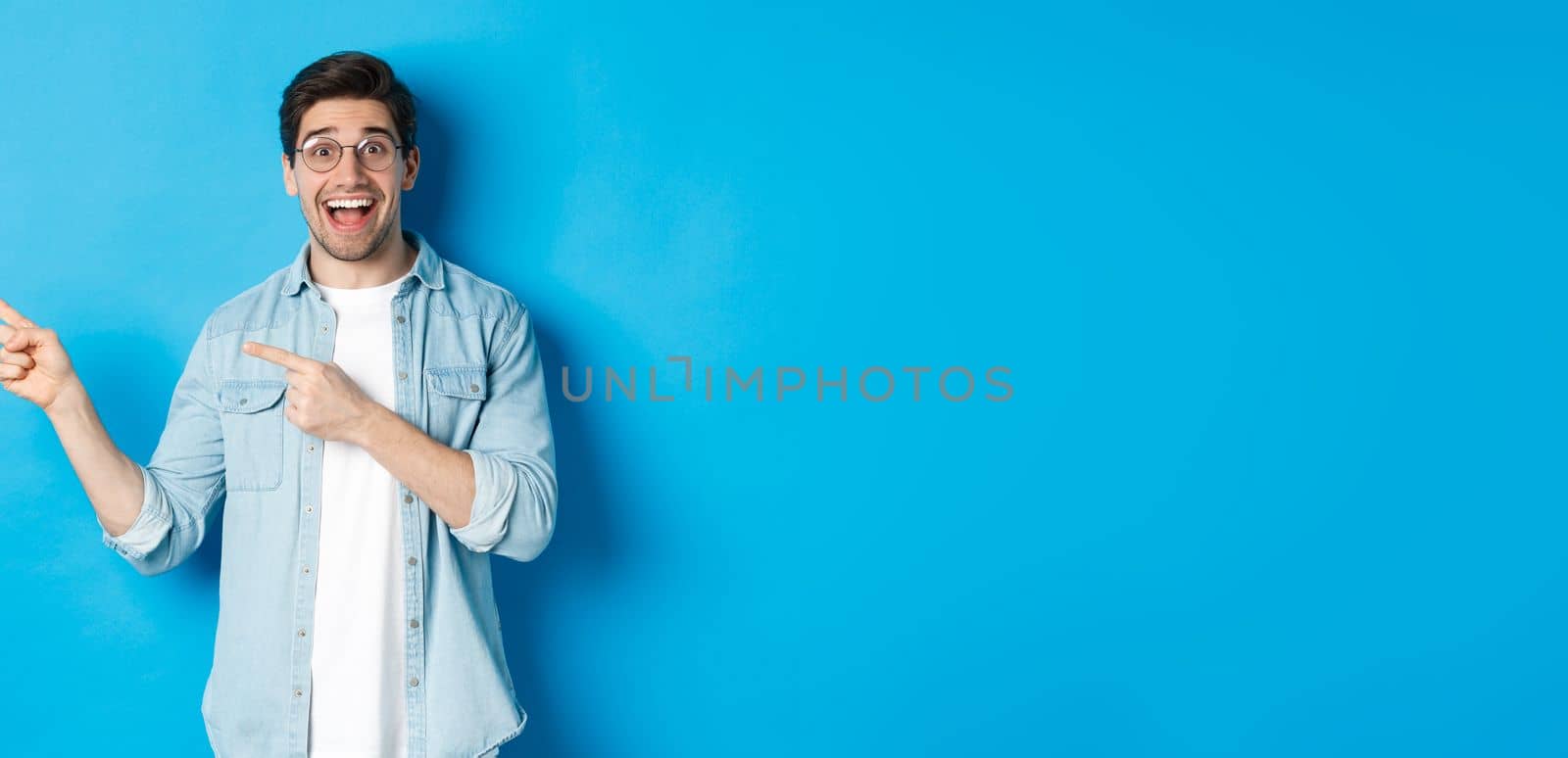 Excited handsome man in glasses pointing fingers left, looking happy while showing advertisement, demonstrating copy space on blue background.