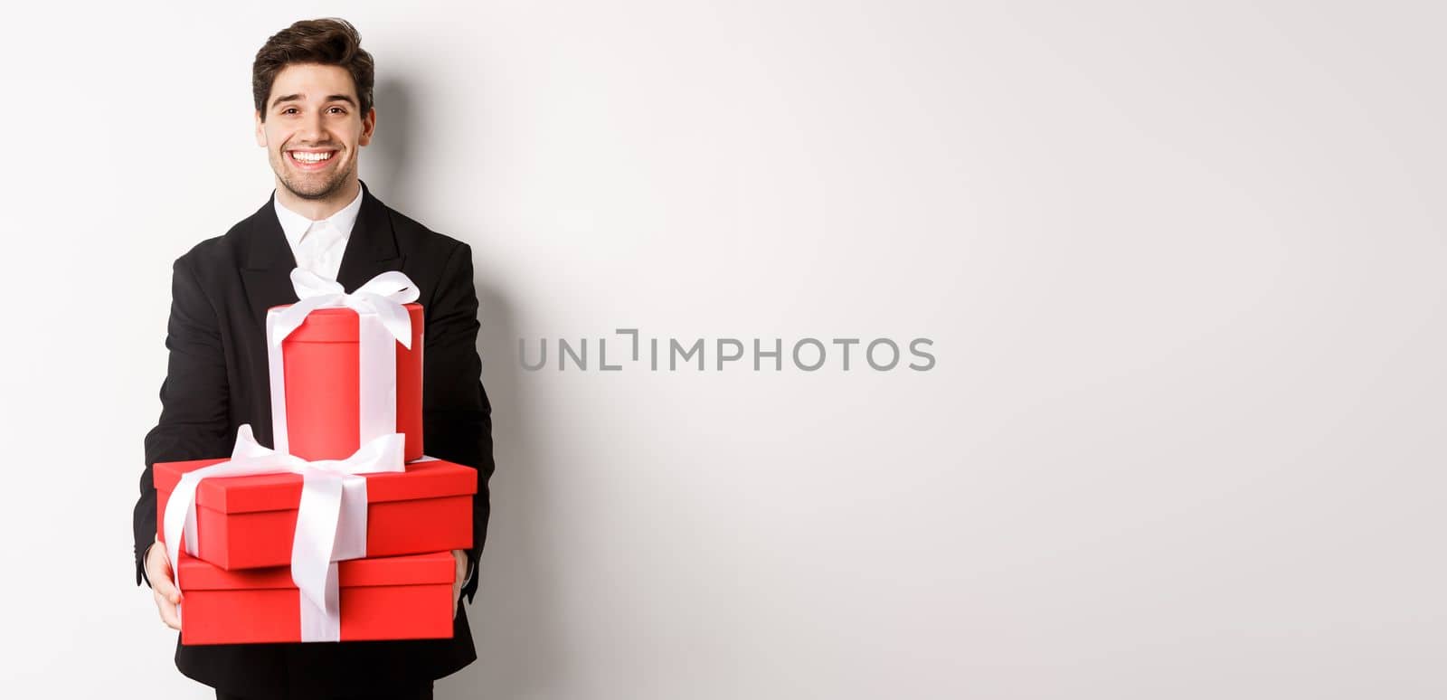 Portrait of handsome bearded man in trendy suit, holding gifts for new year and smiling, prepared presents, standing over white background.