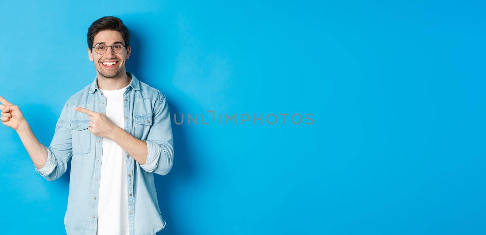 Young handsome man in glasses showing advertisement, smiling and pointing fingers left, making announcement, standing against blue background.