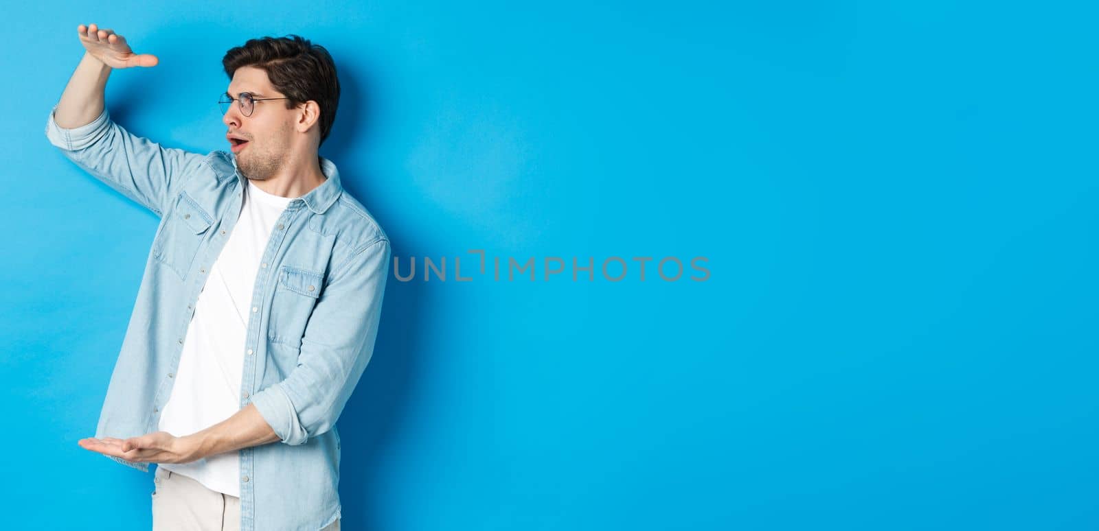 Excited handsome man showing big size object and looking amazed, standing over blue background by Benzoix